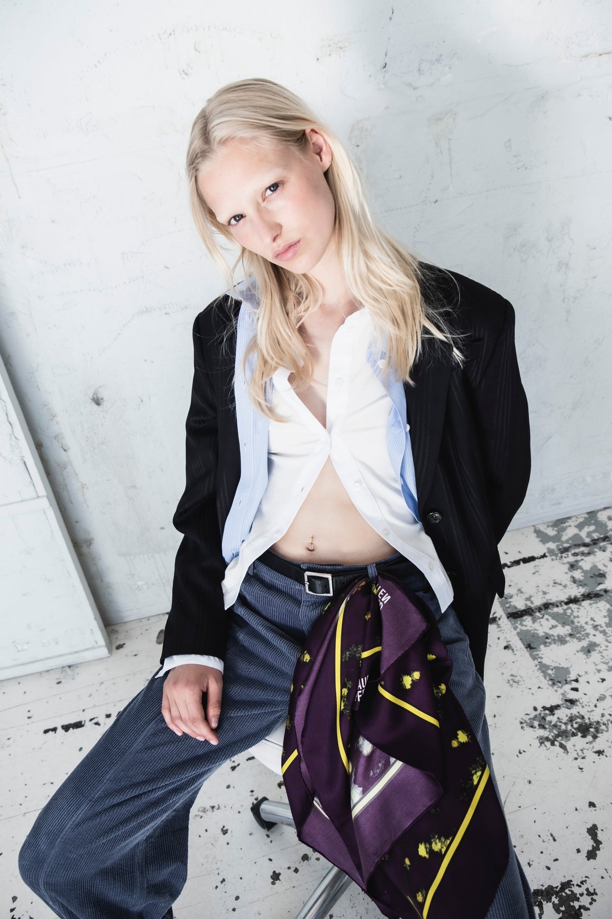 Kampagneportræt af en model i en sort blazer, hvid skjorte med krave og lyseblå pinstribet vest, iført et lilla blomstret tørklæde over mørkeblå bukser.